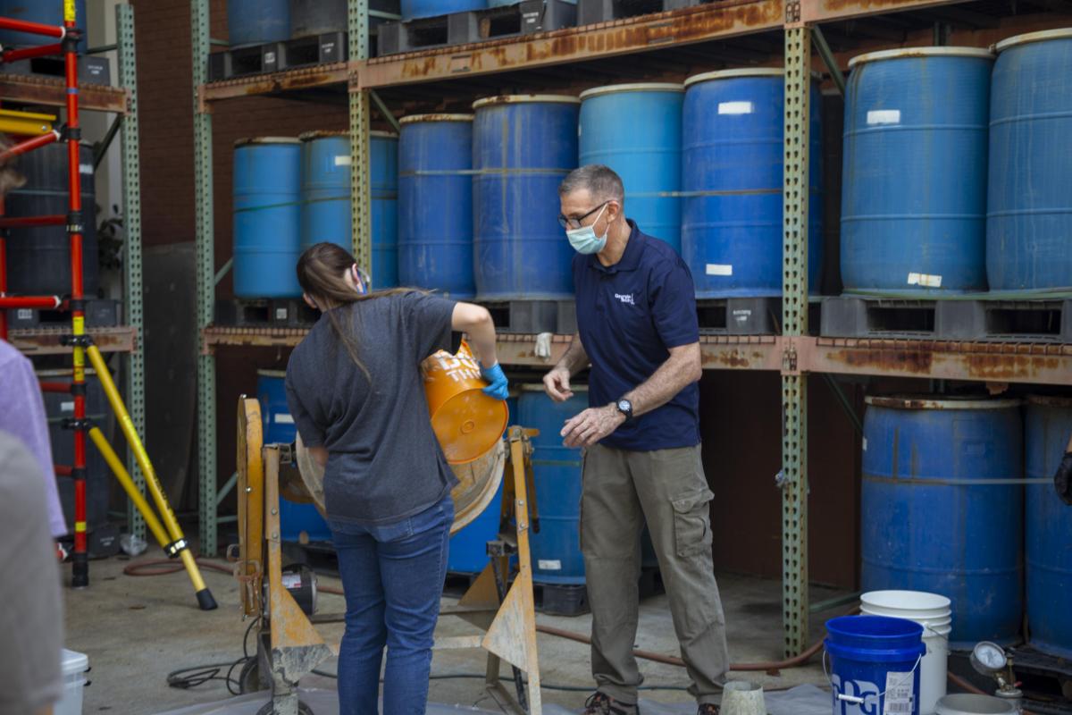 two people mix concrete in front of a shelf of blue barrels
