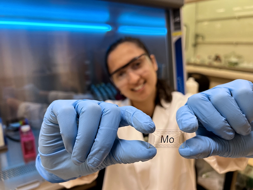 A woman wearing protective glasses and latex gloves holds a glass chip that says "Mo"